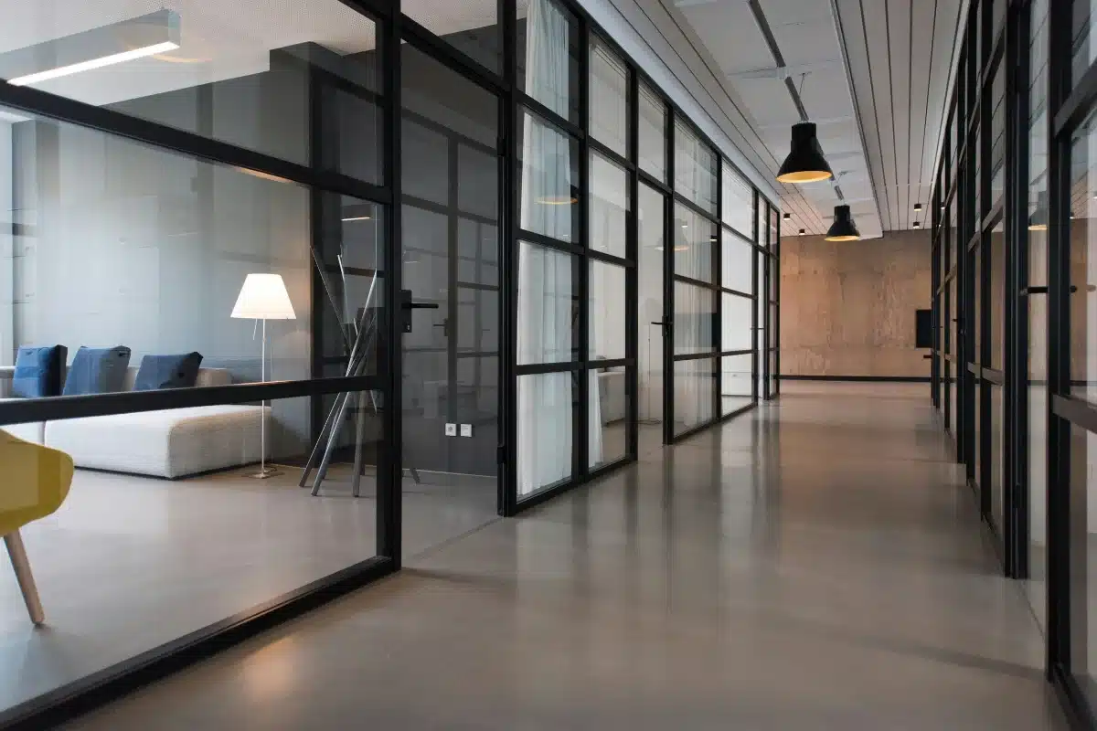 Long glass-walled office corridor with black metal frames and a seating area to the left, including a white sofa and floor lamp.