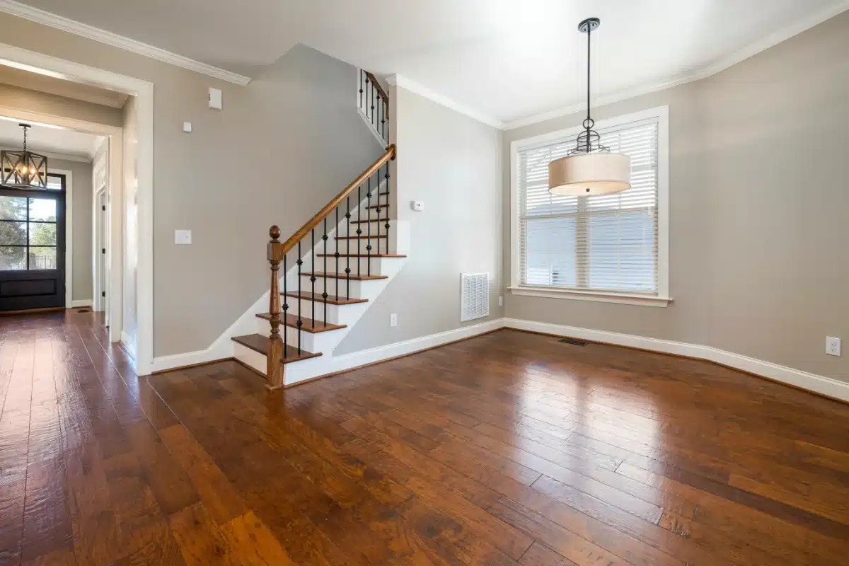 Bright empty living room with hardwood floors, a staircase with wooden railing, and a large window with blinds; contemporary chandelier hangs from the ceiling.